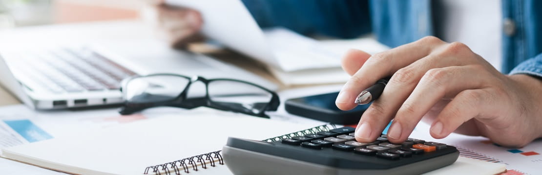 Close-up of a person using a calculator at a desk with documents, a laptop, eyeglasses and a smartphone.