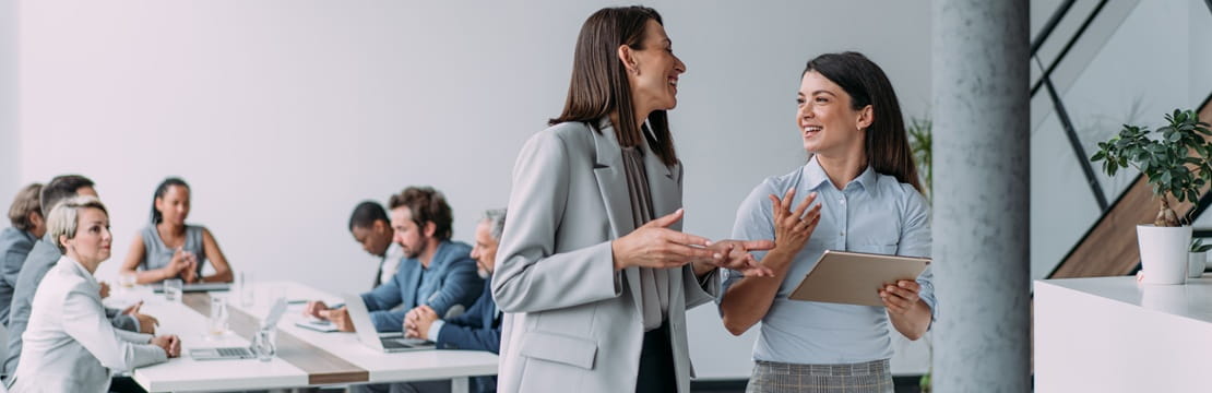 Two colleagues talk while holding a tablet in a modern conference room, with a group of people seated and working at a table in the background.